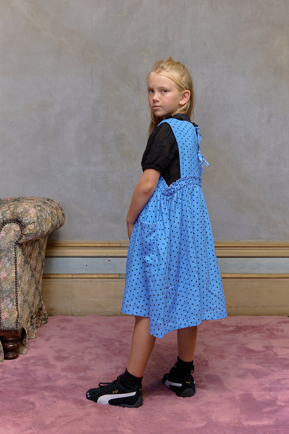 Young girl wearing a blue dress with a black top, standing in a room with pink carpet and floral-patterned chair. Periwinkle blue kids dress in organic cotton muslin with black polka dots and front pockets by Little Harper Lee