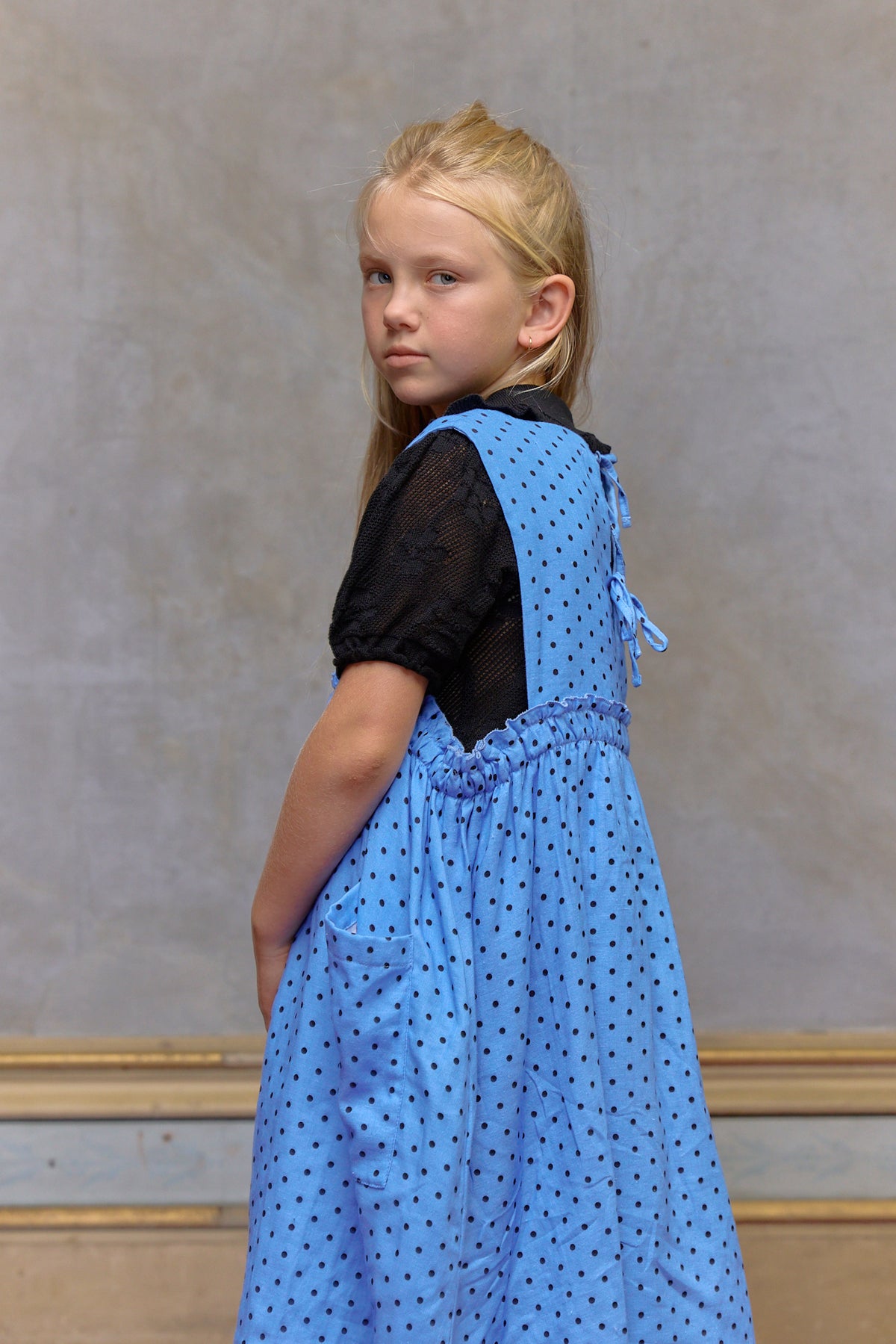 Young girl wearing a blue dress with black polka dots against a plain background. Periwinkle blue kids dress in organic cotton muslin with black polka dots and front pockets by Little Harper Lee