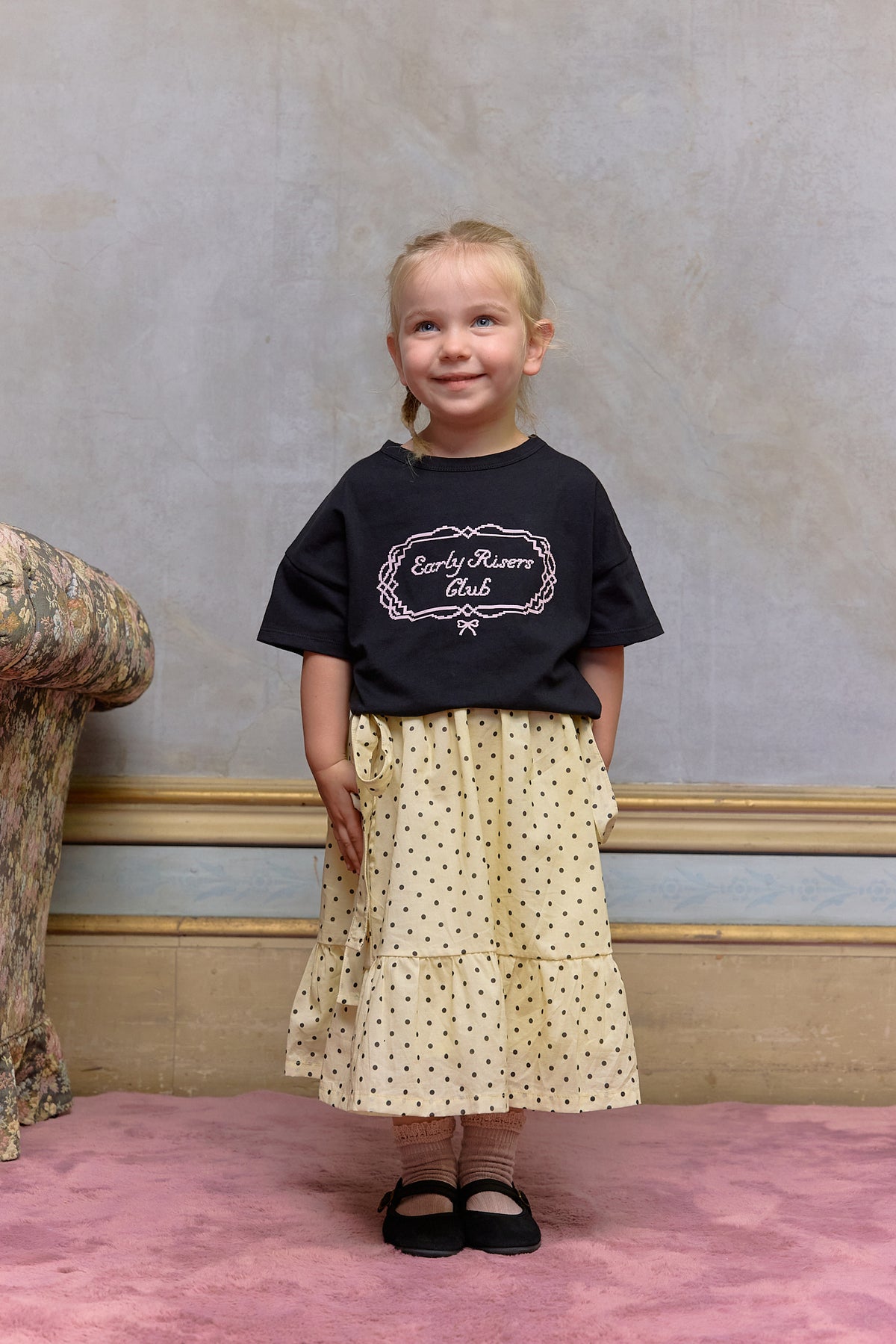 Young girl wearing a black t-shirt with text and a yellow polka dot skirt standing in a room with a floral-patterned chair. Kids cotton graphic t-shirt with Early Risers Club print. Soft, slightly heavier weight fabric with relaxed fit for everyday wear.