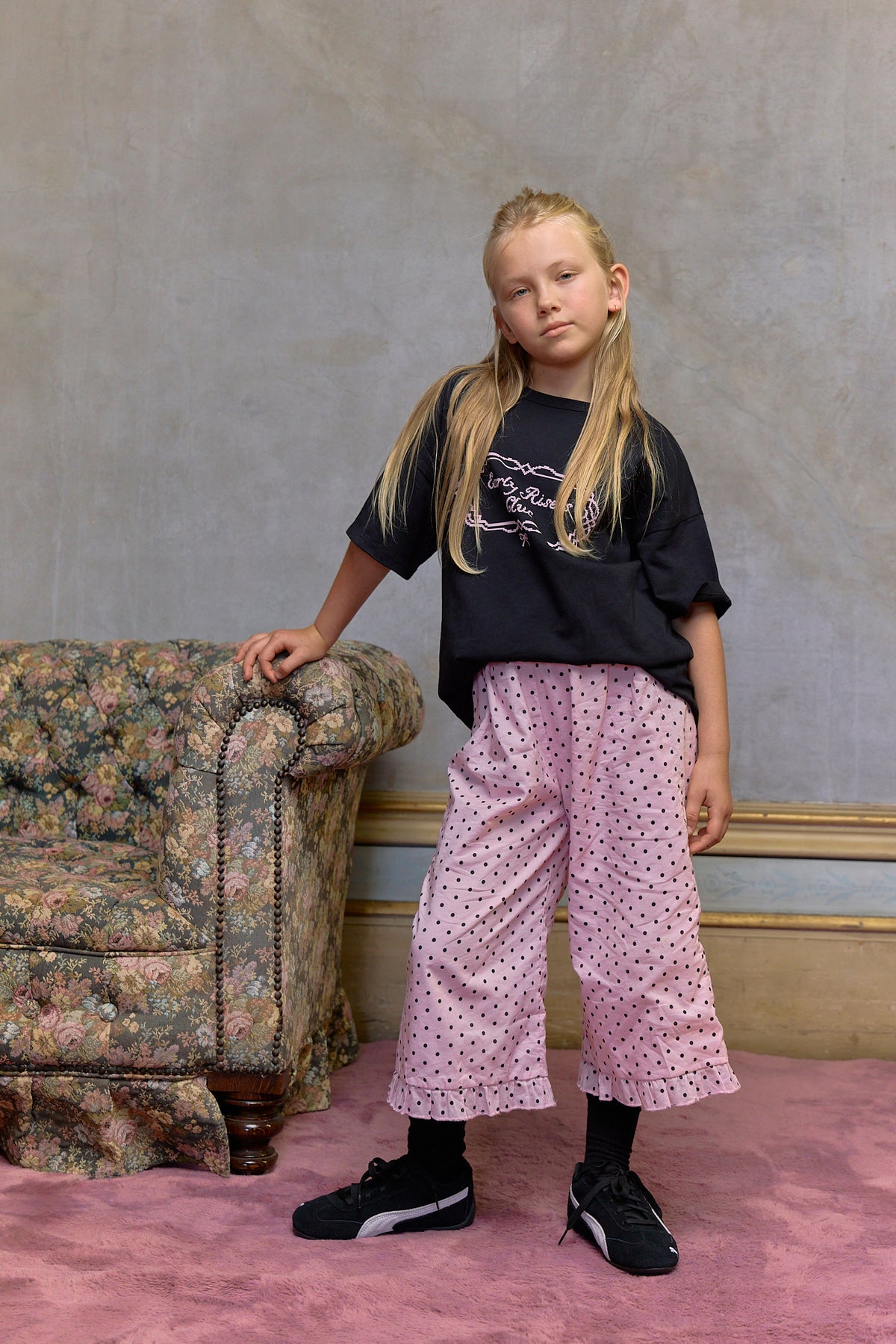 Young girl in a black t-shirt and pink polka dot pants standing in a room with a floral armchair. Kids cotton graphic t-shirt with Early Risers Club print. Soft, slightly heavier weight fabric with relaxed fit for everyday wear.