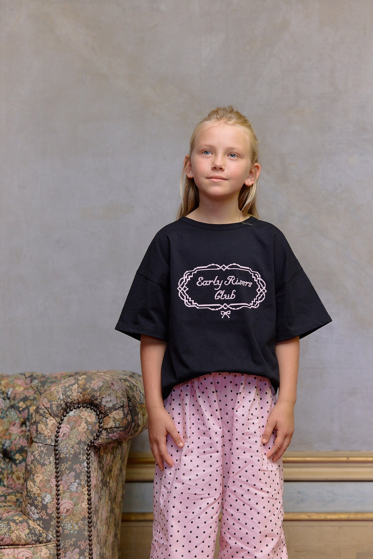 Young girl wearing a black t-shirt with text and pink polka dot pants against a textured wall. Kids cotton graphic t-shirt with Early Risers Club print. Soft, slightly heavier weight fabric with relaxed fit for everyday wear.
