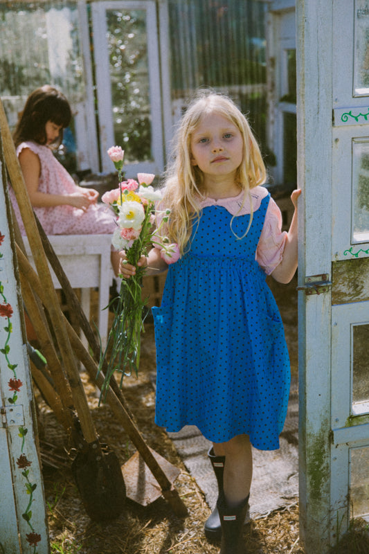 Young girl in a blue dress holding flowers in a garden setting. Periwinkle blue kids dress in organic cotton muslin with black polka dots and front pockets by Little Harper Lee