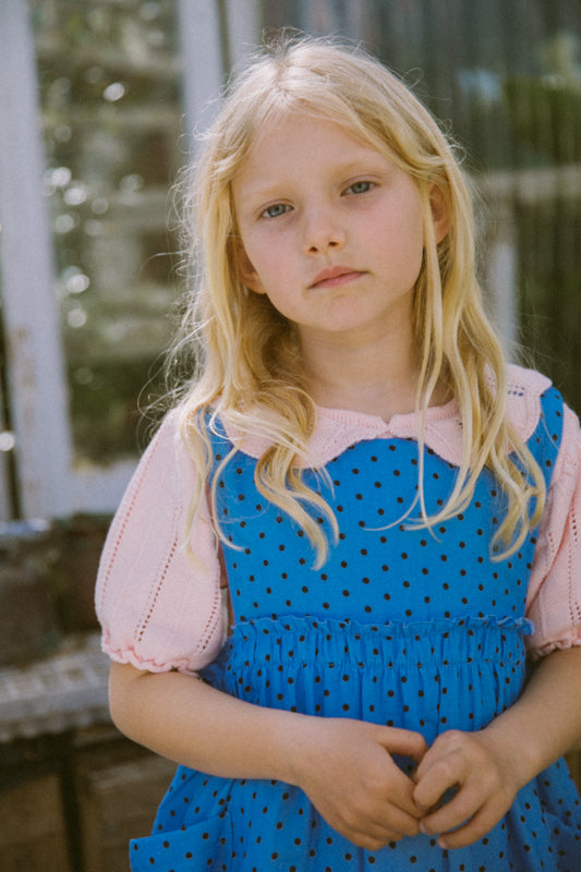 Young girl wearing a blue dress with black polka dots and a pink blouse. Periwinkle blue kids dress in organic cotton muslin with black polka dots and front pockets by Little Harper Lee
