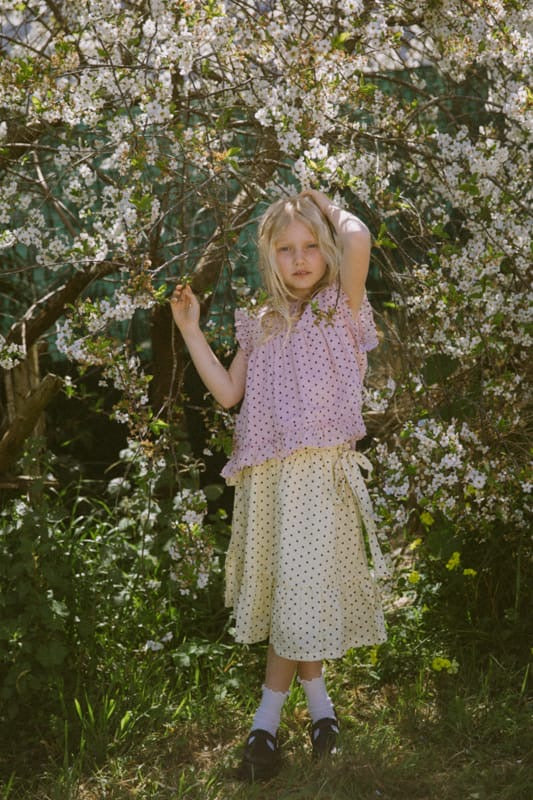 Pink polka dot kids blouse in organic cotton with frill details and button back by Little Harper Lee. standing in front of a cherry blossom tree