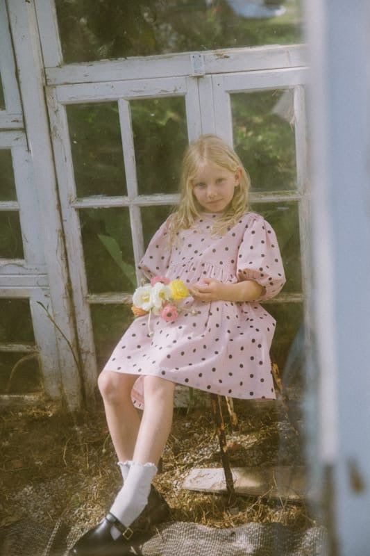 Young girl in a pink polka dot dress holding flowers, sitting on a stone surface with a white window frame in the background.